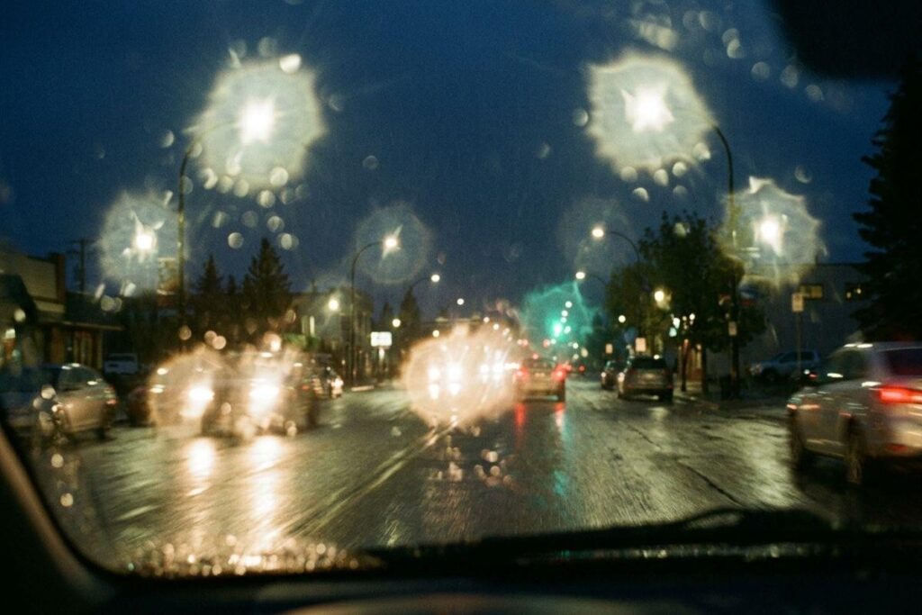 Rainy night street seen through a rain-covered windshield, with blurred street lights, wet road reflections, and oncoming car headlights.